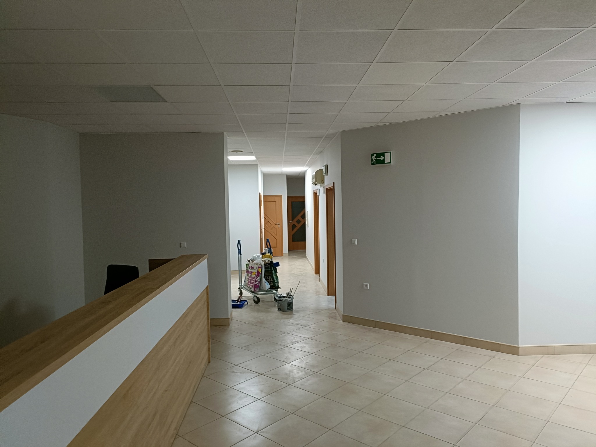 Korytarz biurowy z recepcją, świeżo po malowaniu ścian na jasny odcień szarości, w tle wózek z narzędziami malarskimi i wiaderkiem farby.