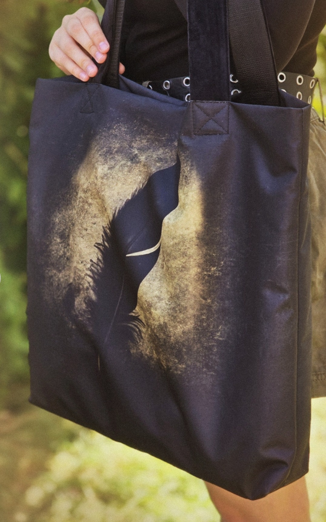 Czarna torba na ramię z nadrukiem pióra, trzymana przez osobę z widocznym paskiem z kółkami, na rozmytym zielonym tle.