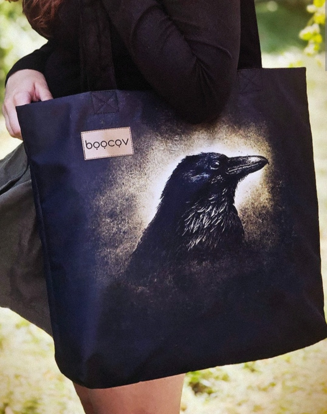 Czarna torba na ramię z nadrukiem kruka i skórzaną naszywką z logo, trzymana przez osobę na tle zieleni.