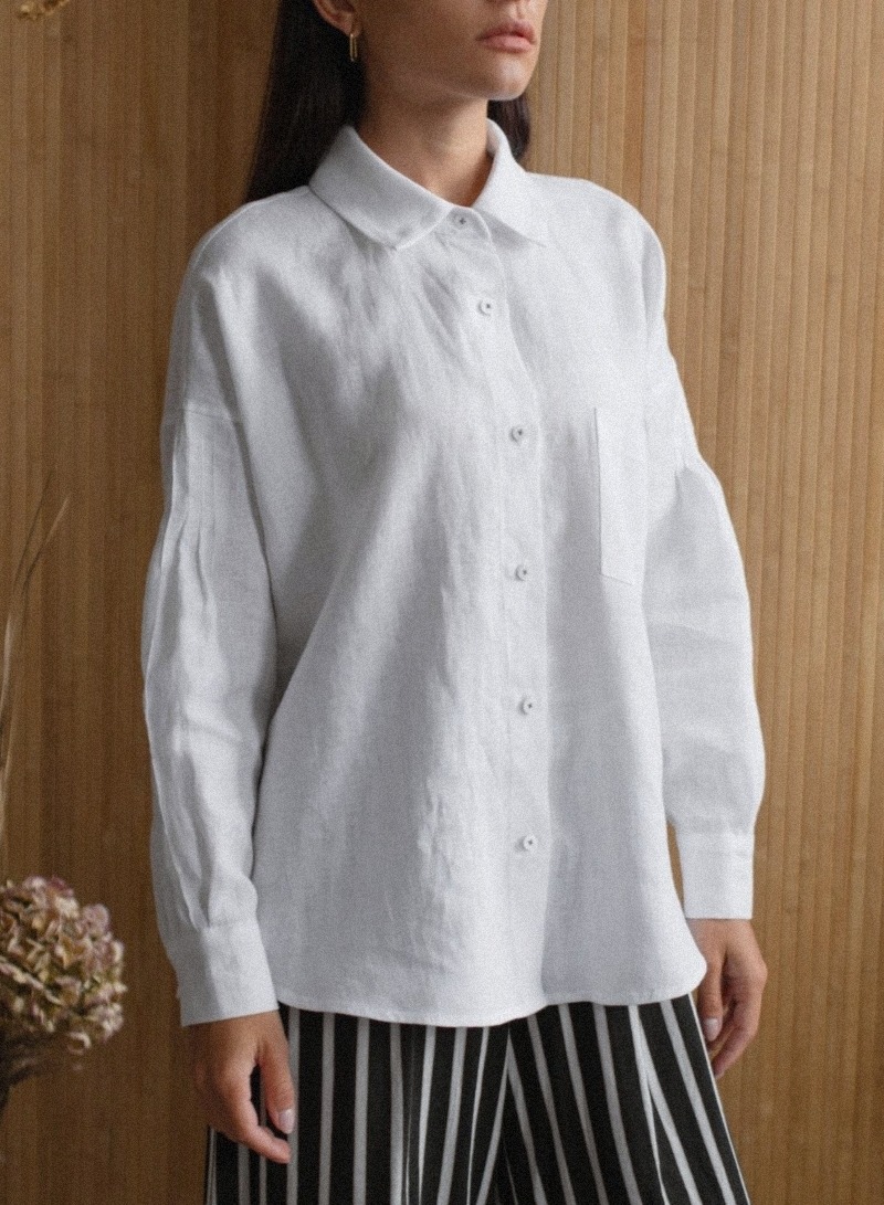 Prezentacja damskiej, białej, lnianej koszuli z długim rękawem, z kieszenią na piersi, zestawionej z spodniami w czarno-białe pionowe pasy, na tle drewnianej ściany.