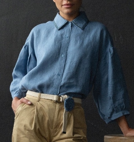 Kobieta w błękitnej lnianej koszuli z haftowanym kołnierzykiem i beżowych spodniach z plecionym paskiem, oparta o drewniany stołek na ciemnym tle.