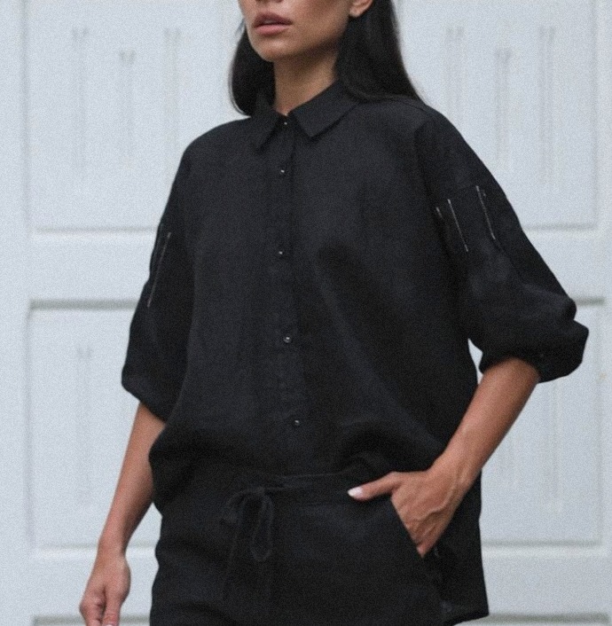 Modelka prezentuje czarny, lniany komplet: koszula z krótkim rękawem z ozdobnymi szwami i spodnie ze sznurkiem w pasie na tle białych drzwi.