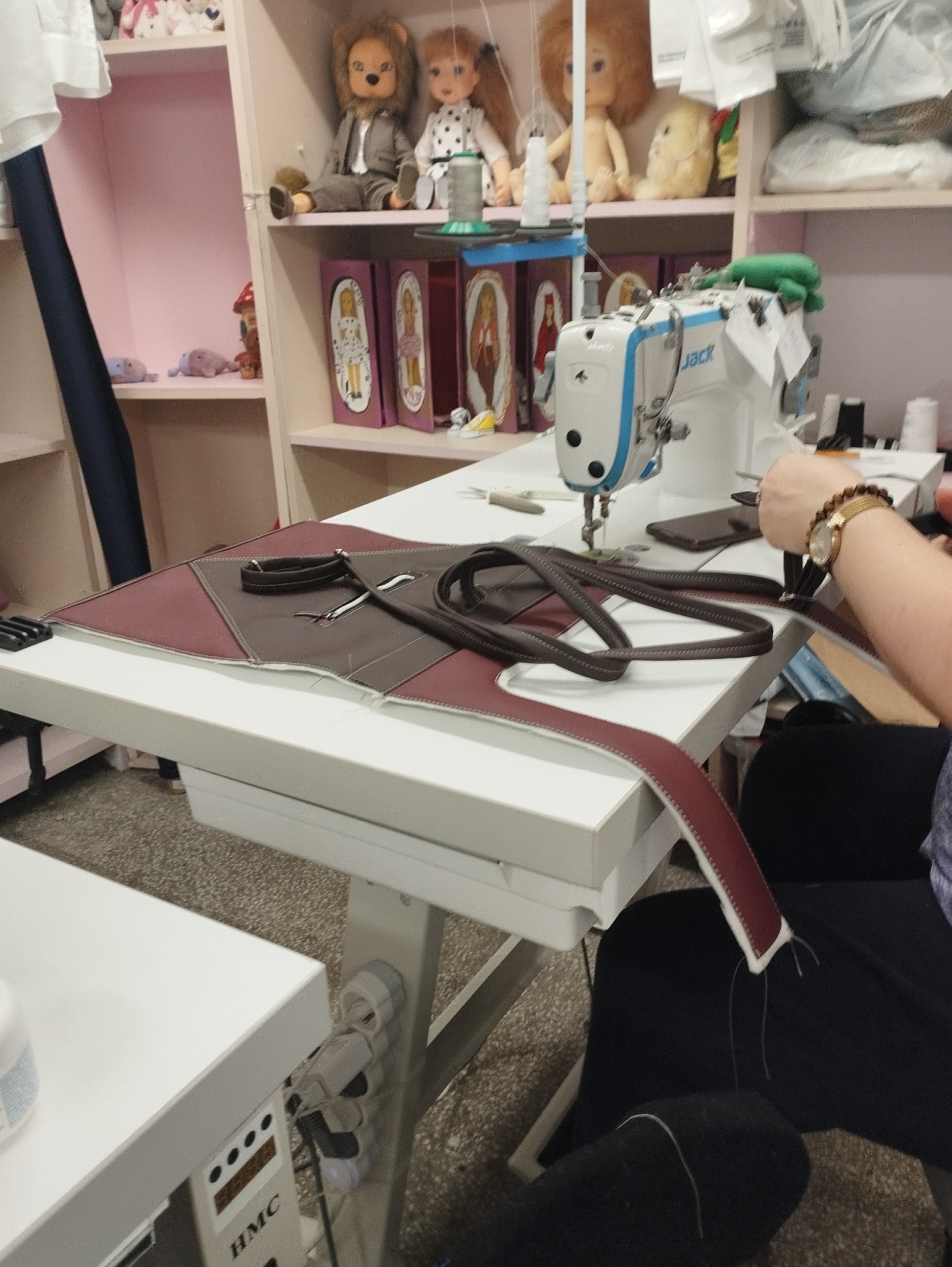 Praca przy szyciu skórzanej torby na maszynie szwalniczej, widoczna ręka z zegarkiem, w tle półki z dekoracjami i zabawkami.