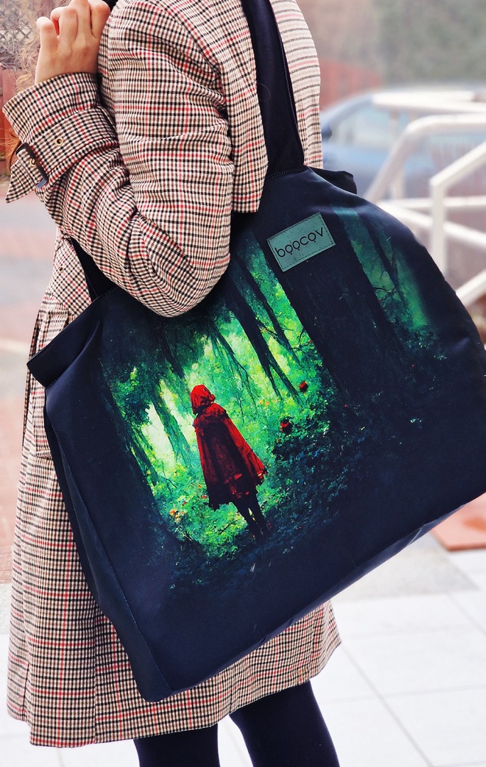 Torba shopper z nadrukiem Czerwonego Kapturka w lesie, noszona na ramieniu przez osobę w płaszczu w kratę, widoczne logo marki na małej naszywce.