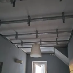 Metalowa konstrukcja sufitu podwieszanego z płyty gipsowo-kartonowej, widoczne profile i zawiesia, w tle szara ściana z oknem i lampą.