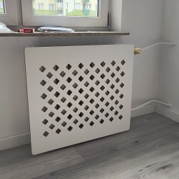 DOBPOL - Biała, ażurowa osłona grzejnika w rogu pokoju z szarą podłogą i białymi ścianami. Widoczne rury grzewcze i parapet z akcesoriami.