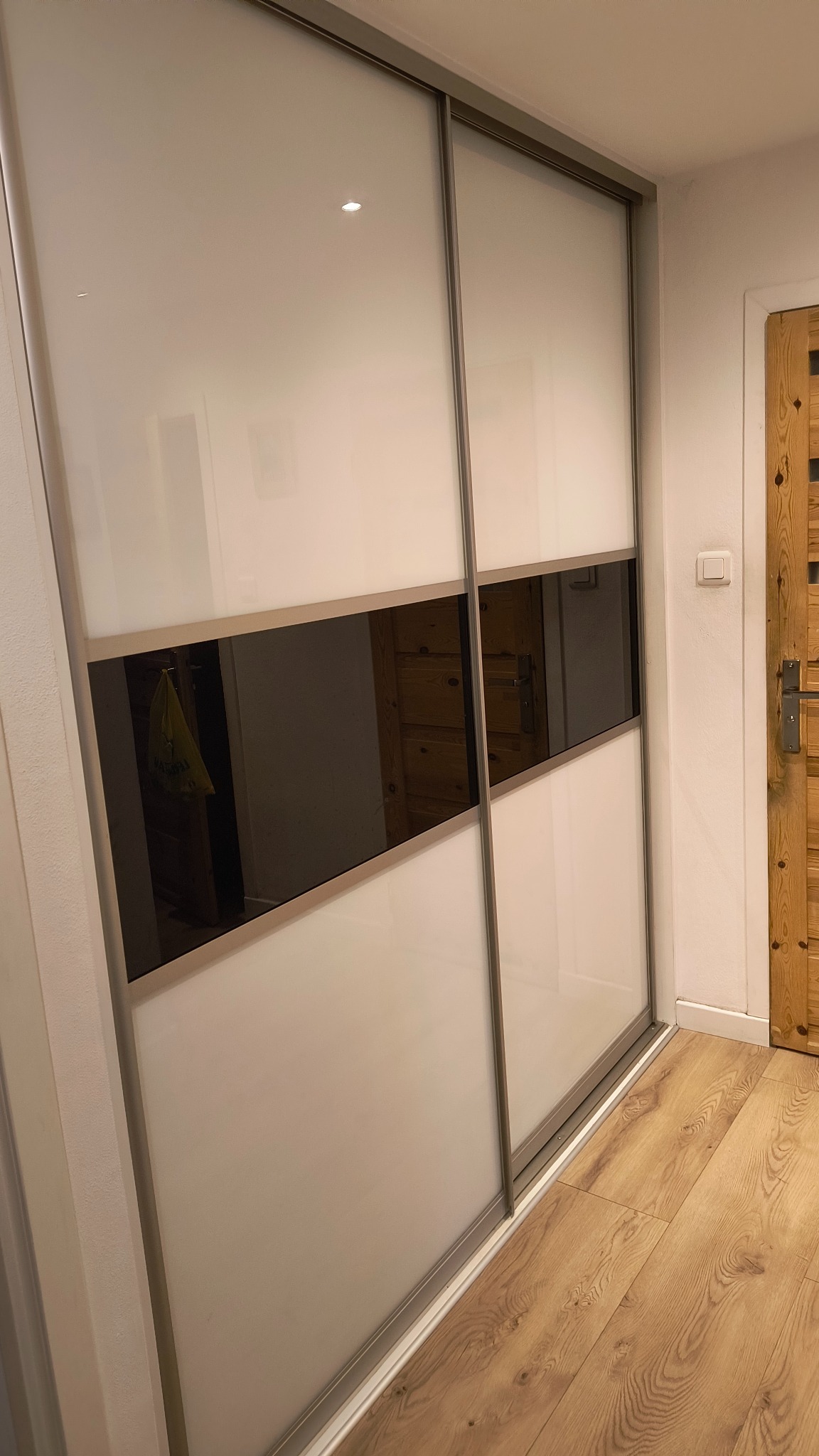 Biała szafa przesuwna z czarnym pasem pośrodku, odbijająca drzwi z jasnego drewna i włącznik światła.