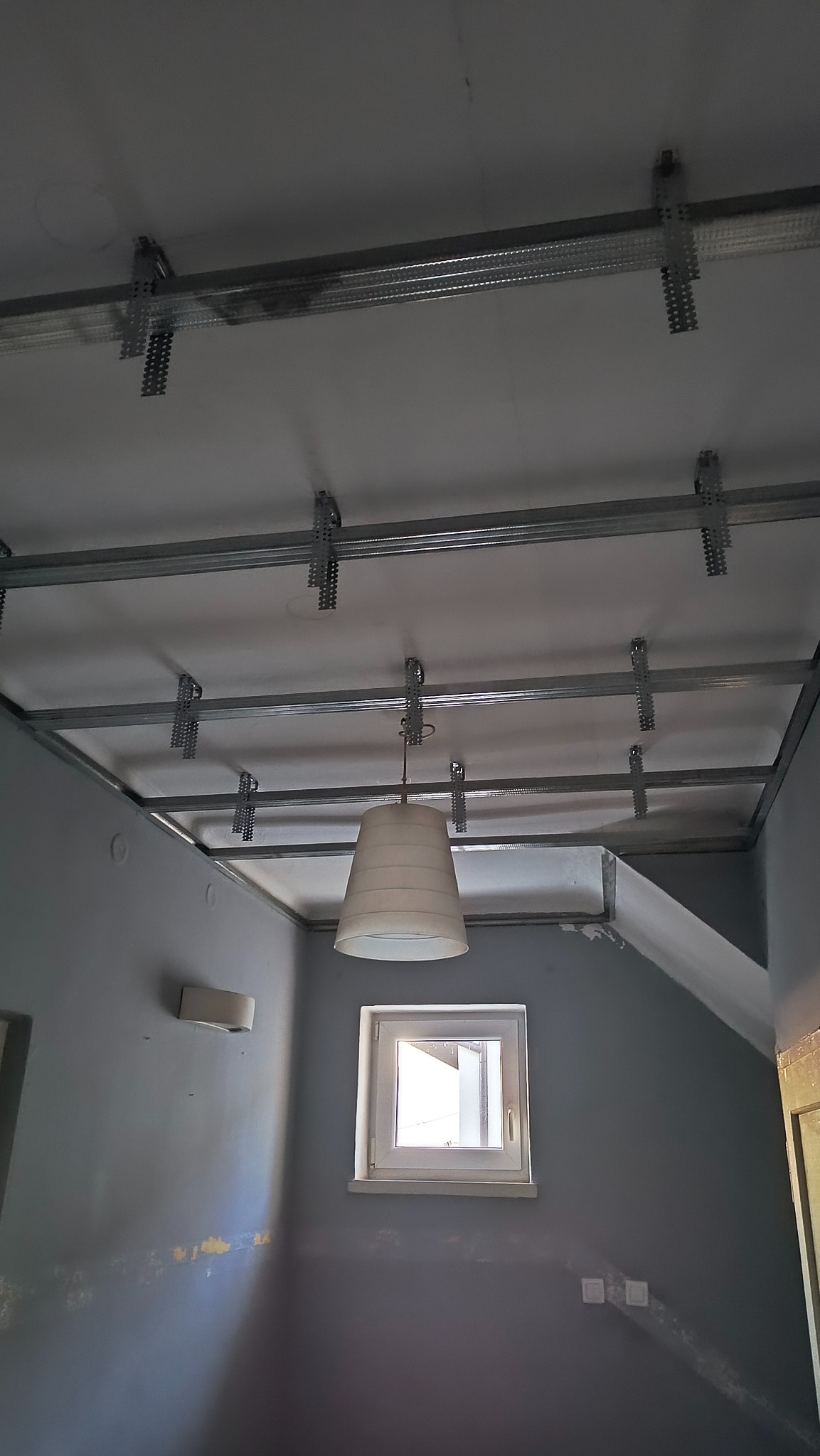Metalowa konstrukcja sufitu podwieszanego z płyty gipsowo-kartonowej, widoczne profile i zawiesia, w tle szara ściana z oknem i lampą.