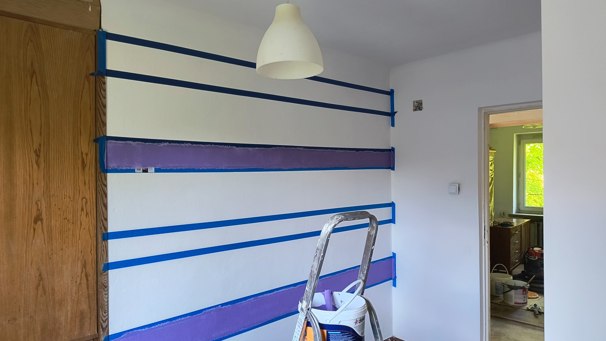 Ściana w pokoju z poziomymi pasami malowanymi na fioletowo, oddzielonymi niebieską taśmą malarską. Widoczna drabina i wiadro z farbą. W tle otwarte drzwi.