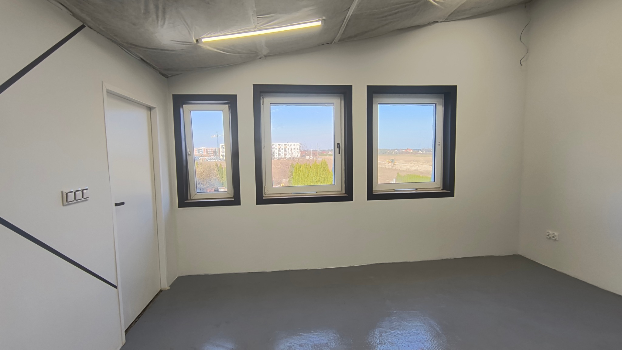 Wnętrze pokoju po remoncie: szara podłoga, białe ściany z czarnym akcentem, trzy okna w ciemnych ramach. Widok na zewnątrz z budynkami i zielenią.