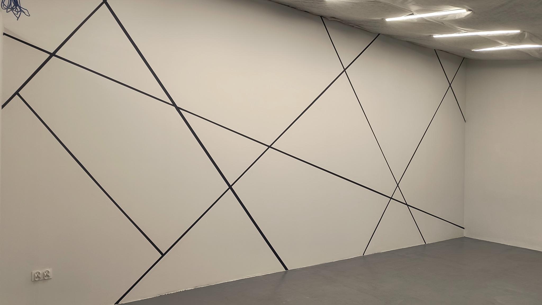 Biała ściana z dekoracyjnymi czarnymi liniami tworzącymi geometryczny wzór. Szare podłoże, oświetlenie sufitowe.
