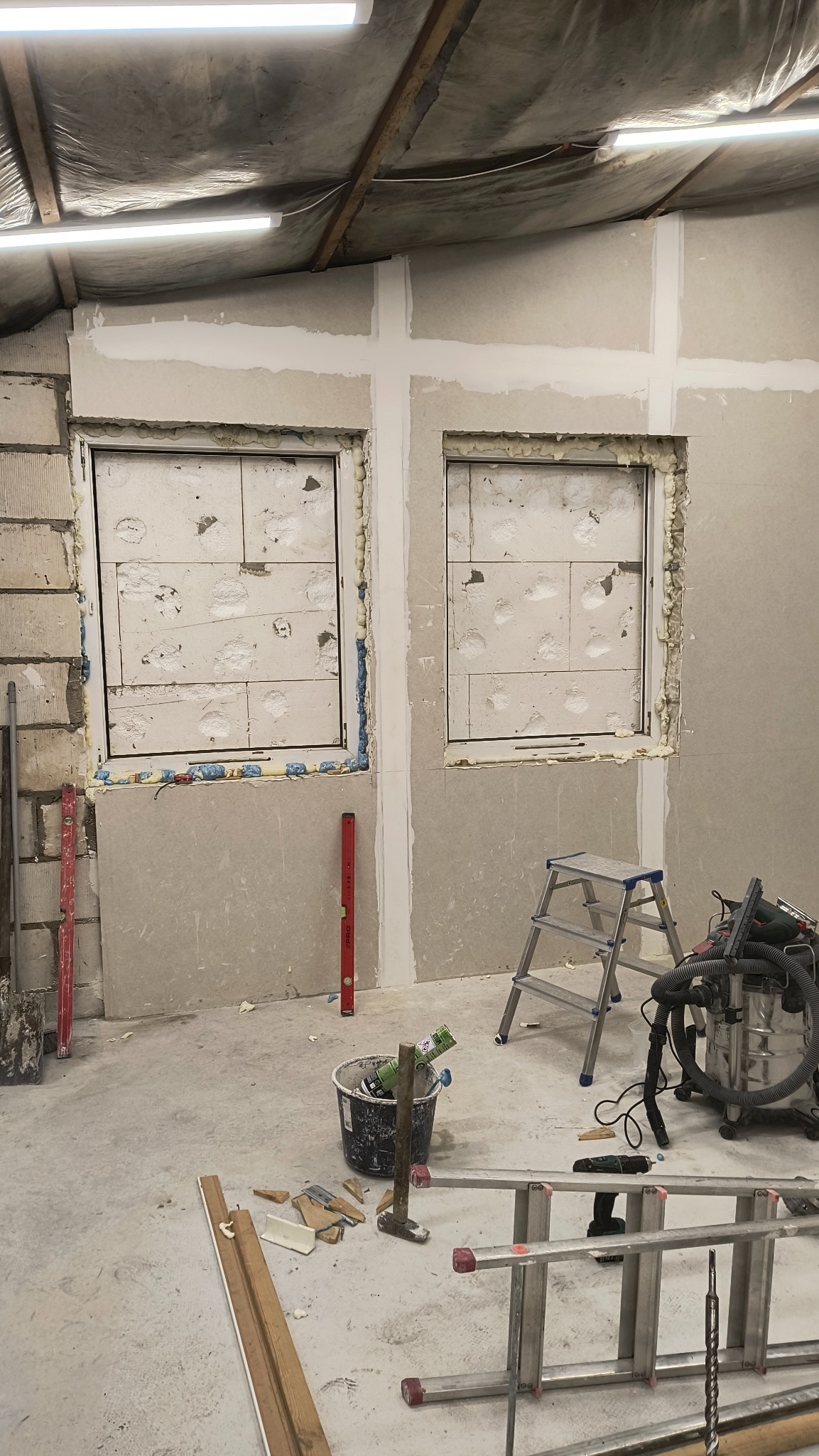 Montaż płyt gipsowo-kartonowych wokół okien, widoczne narzędzia i drabina. Etap prac przygotowawczych w pomieszczeniu z surowym oświetleniem.