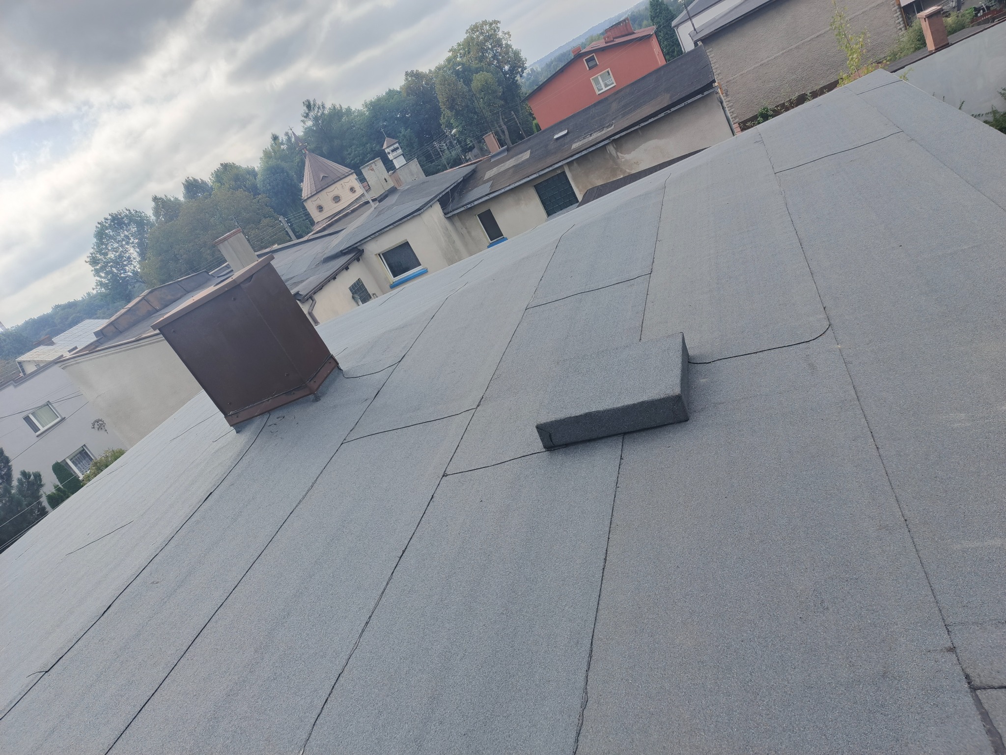 Szary, płaski dach pokryty papą termozgrzewalną z kominem wentylacyjnym i fragmentem attyki. W tle zabudowa miejska i zieleń. Ujęcie z góry, lekko z ukosa.