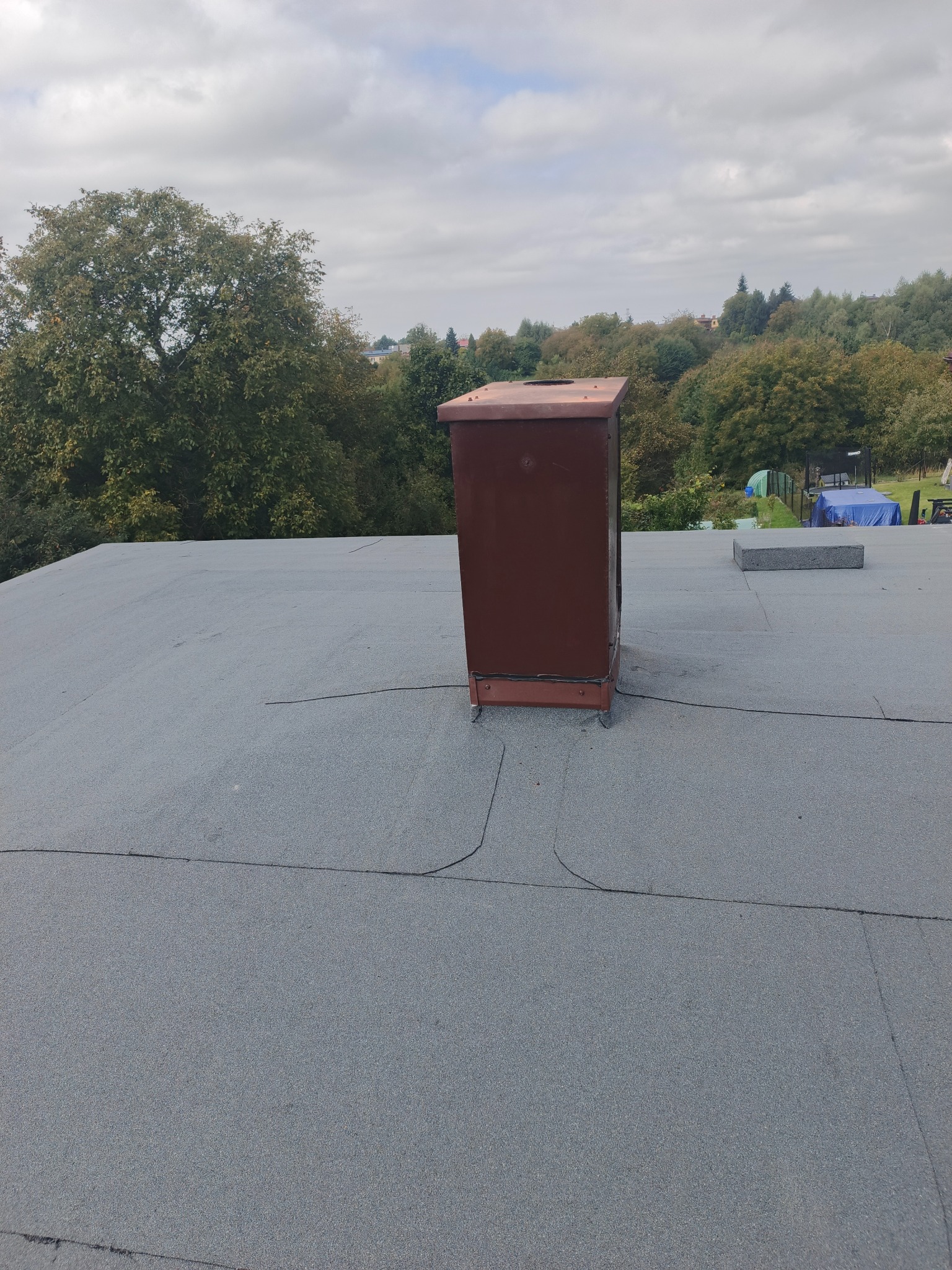 Szary dach pokryty papą z brązowym kominem wentylacyjnym na tle zieleni i pochmurnego nieba. Widoczne łączenia papy. Kąt ujęcia z góry.