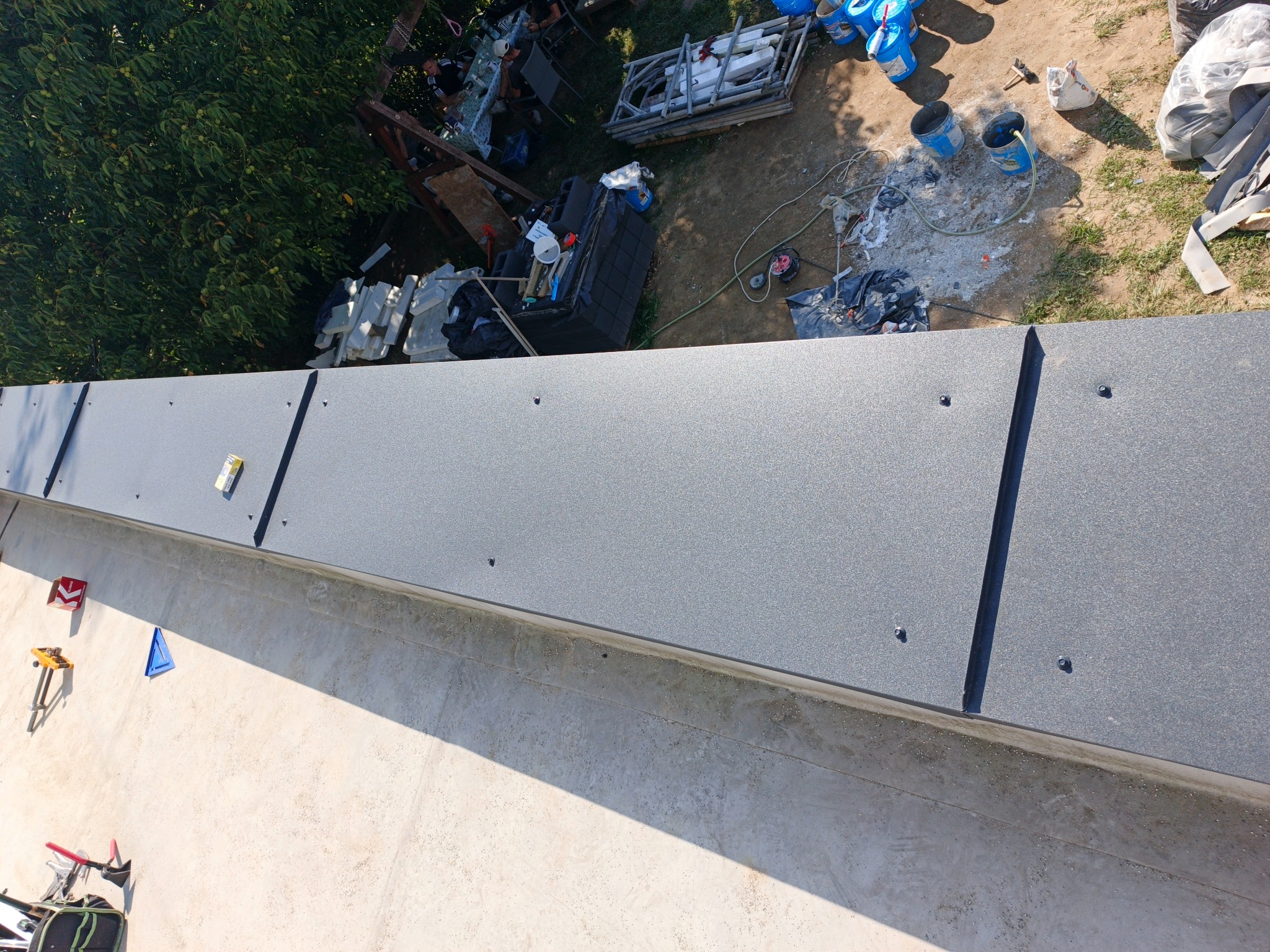 Szary, metalowy panel dachowy z widocznymi mocowaniami, świeżo zamontowany na dachu. W tle narzędzia i materiały budowlane, sugerujące trwające prace dekarskie.