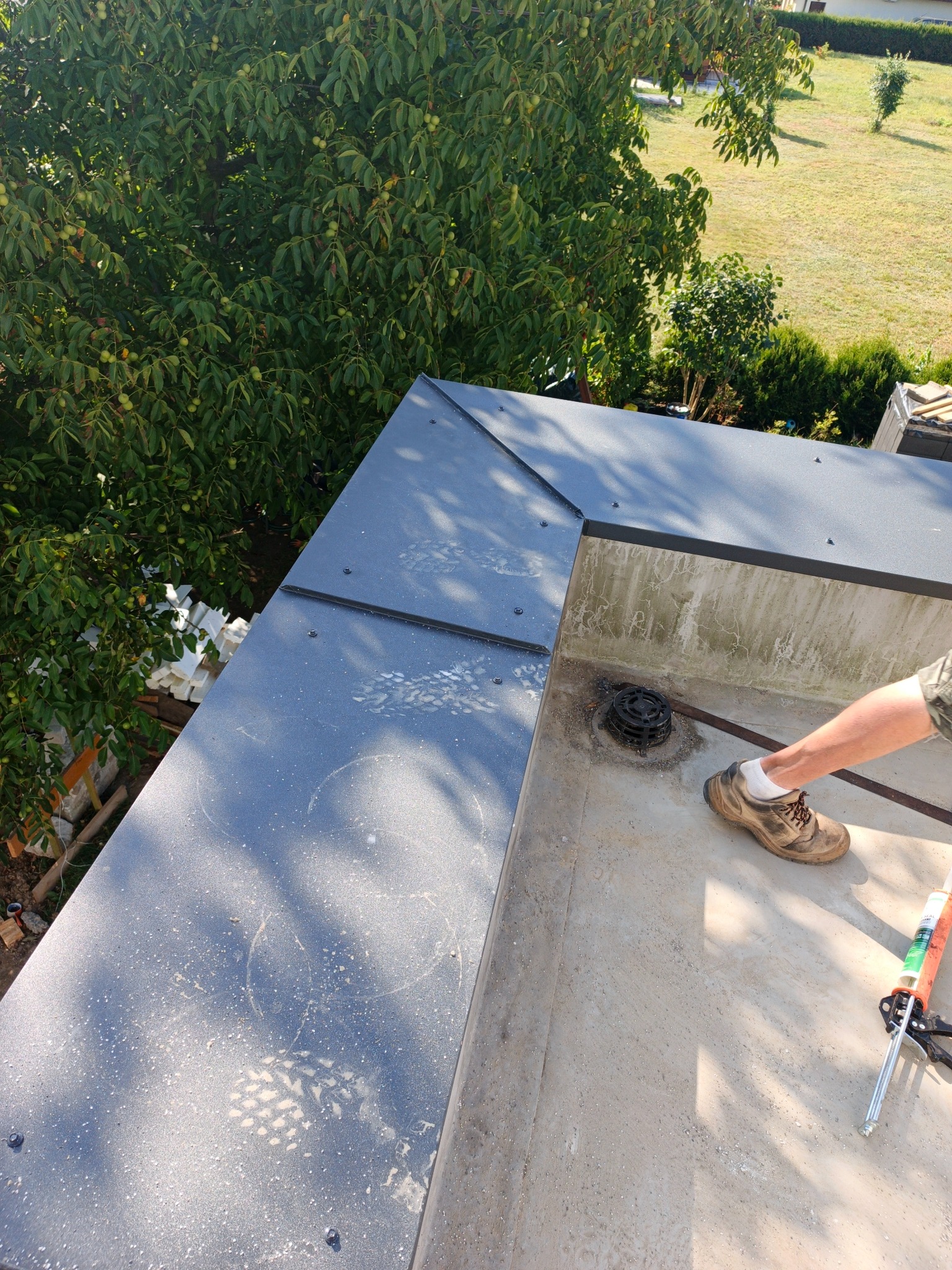 Szary dach płaski z widocznym fragmentem nogi dekarza, narzędziami i elementami wykończeniowymi. W tle zieleń drzew i trawnik. Widoczne ślady na powierzchni dachu.