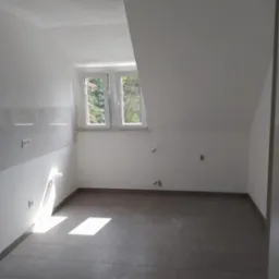 Puste pomieszczenie na poddaszu po remoncie, z ułożonymi szarymi płytkami na podłodze, świeżo pomalowanymi białymi ścianami, widocznymi gniazdkami elektrycznymi i wyprowadzonymi instalacjami...