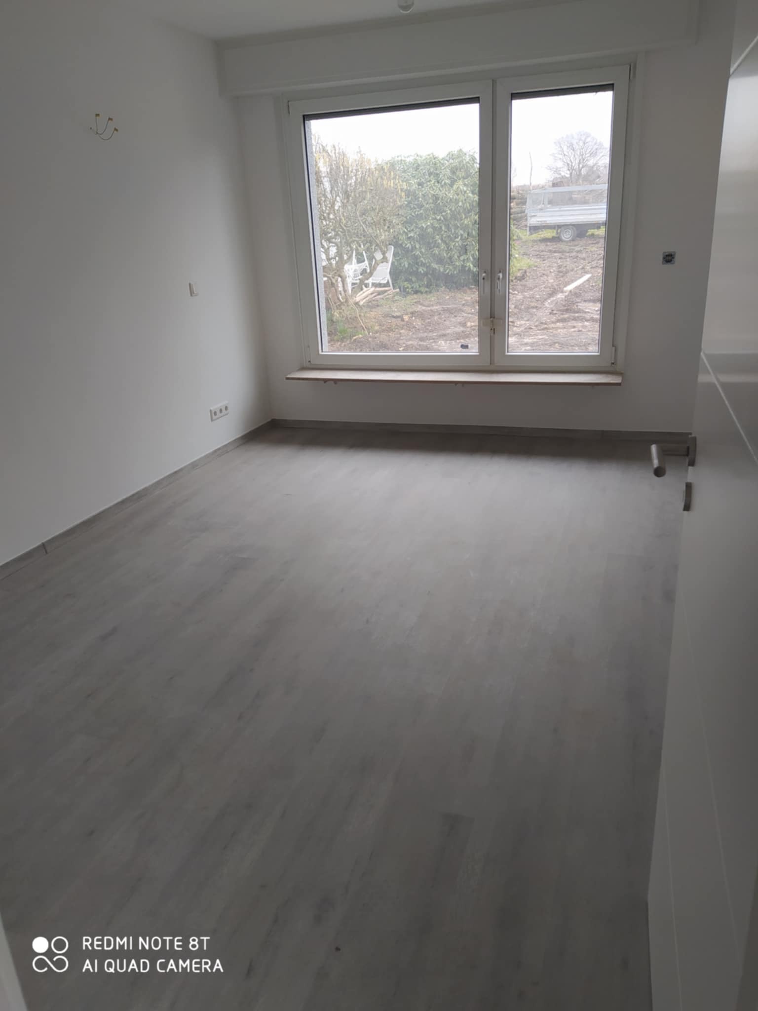 Pusty pokój po remoncie z szarą podłogą imitującą drewno, białe ściany, okno z widokiem na ogród i przyczepę.