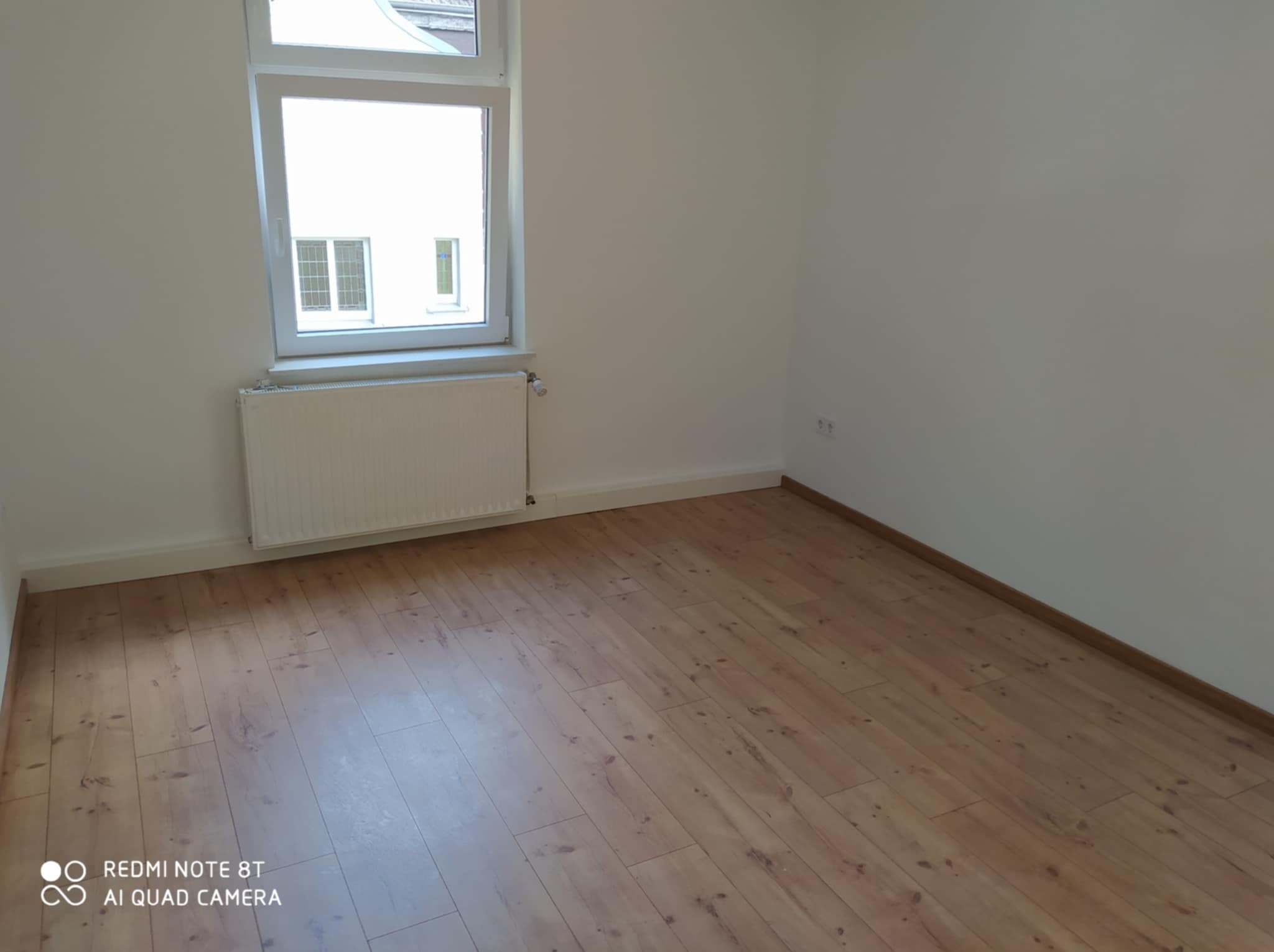 Pusty pokój po remoncie z jasnymi ścianami, panelami podłogowymi w odcieniach brązu i białym grzejnikiem pod oknem.