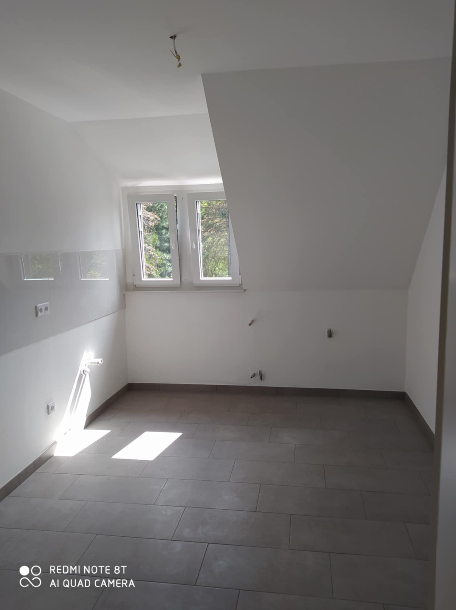 Puste pomieszczenie na poddaszu po remoncie, z ułożonymi szarymi płytkami na podłodze, świeżo pomalowanymi białymi ścianami, widocznymi gniazdkami elektrycznymi i wyprowadzonymi instalacjami...