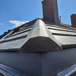 Creativ Dach Artur Klusek - Nowe pokrycie dachu z blachy na rąbek stojący, widok z dołu na krawędź dachu, rynny i komin na tle błękitnego nieba.