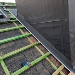 Creativ Dach Artur Klusek - Montaż obróbek blacharskich komina na dachu z membraną Therm, widoczne zielone drewniane elementy konstrukcji dachu i robocze rękawice dekarza.