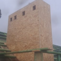 Creativ Dach Artur Klusek - Konstrukcja komina z płyt OSB z czterema otworami wentylacyjnymi w trakcie budowy, widoczny dekarz na dachu z zielonymi łatami w pochmurny dzień.