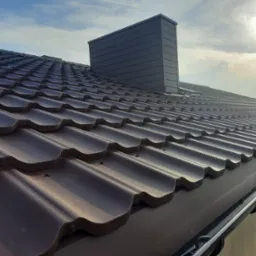 Nowy, brązowy dach z blachodachówki modułowej, z widocznym kominem w kolorze grafitowym, zamontowany na budynku z rynną w kolorze dachu, sfotografowany w słoneczny dzień.