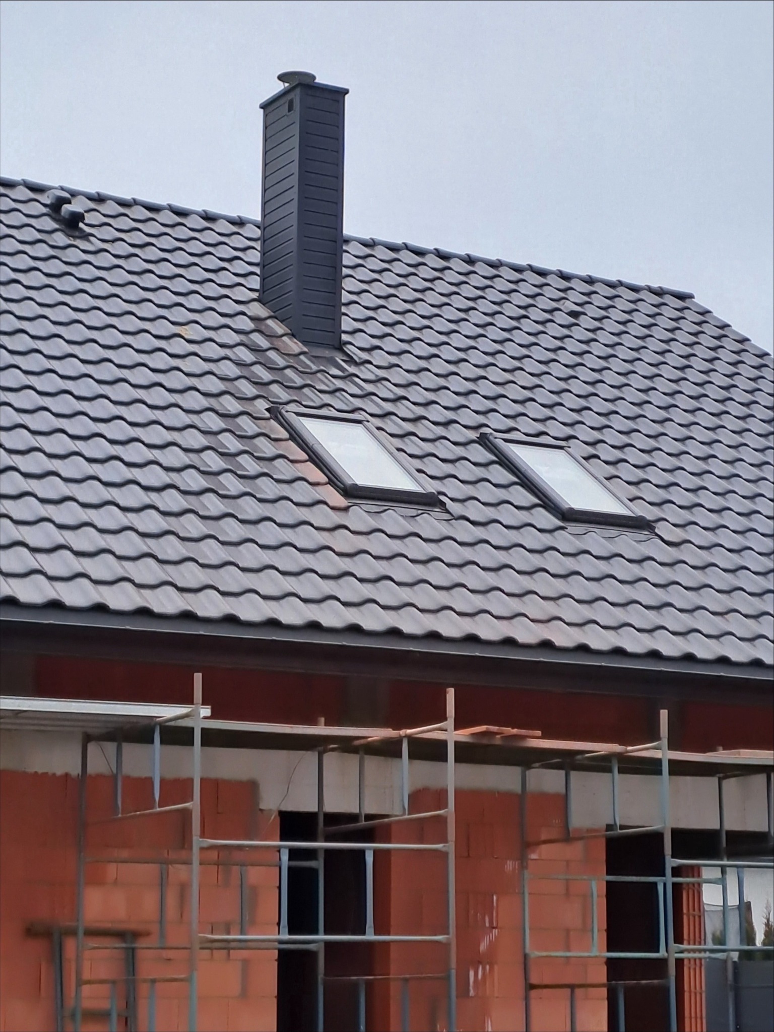 Szary dach z dachówki ceramicznej z dwoma oknami dachowymi i kominem wentylacyjnym na tle szarego nieba, widoczne rusztowania na budynku w trakcie budowy.