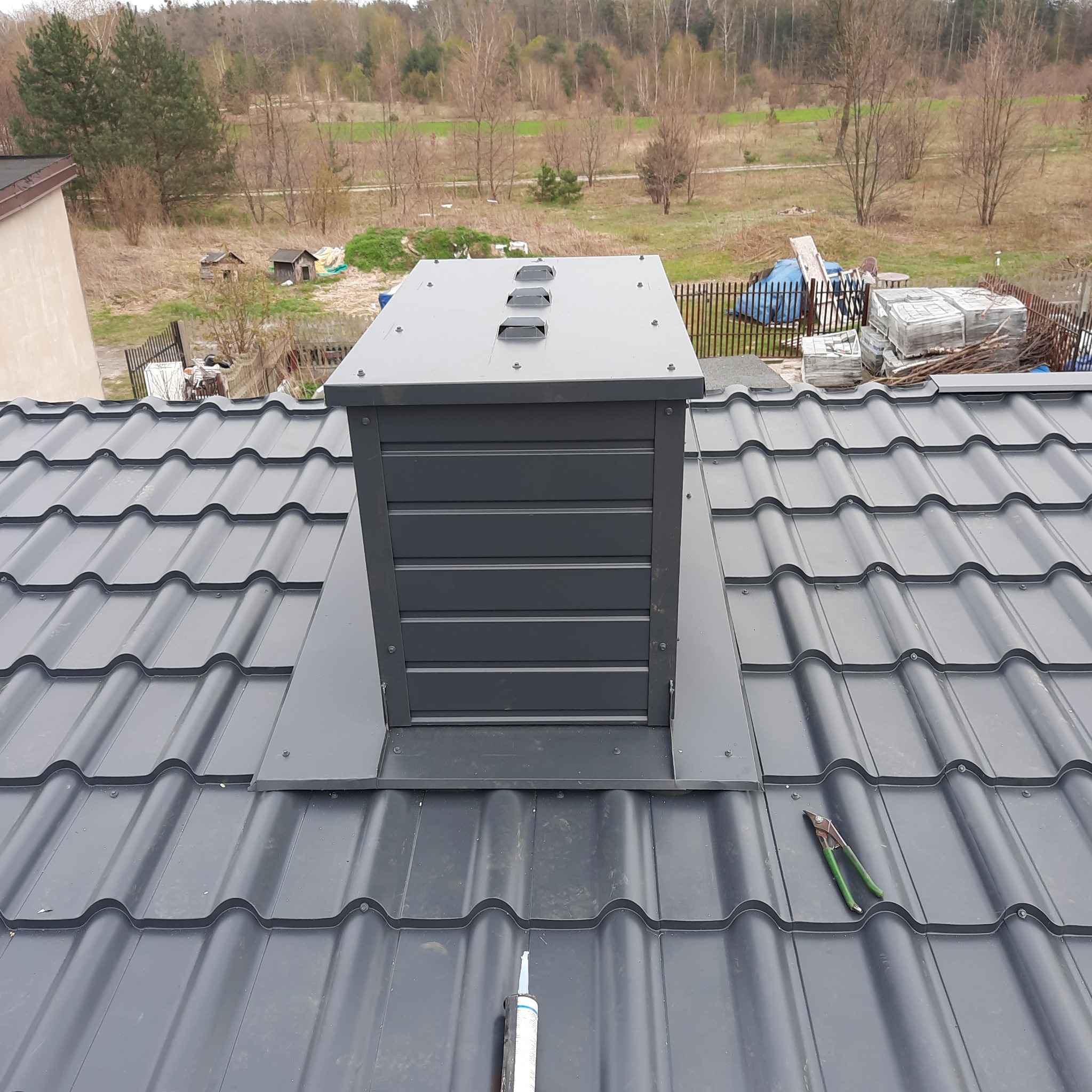 Nowoczesny, grafitowy komin wentylacyjny zamontowany na dachu z blachodachówki, widoczne narzędzia i tuba silikonu dekarskiego, w tle zabudowa jednorodzinna.