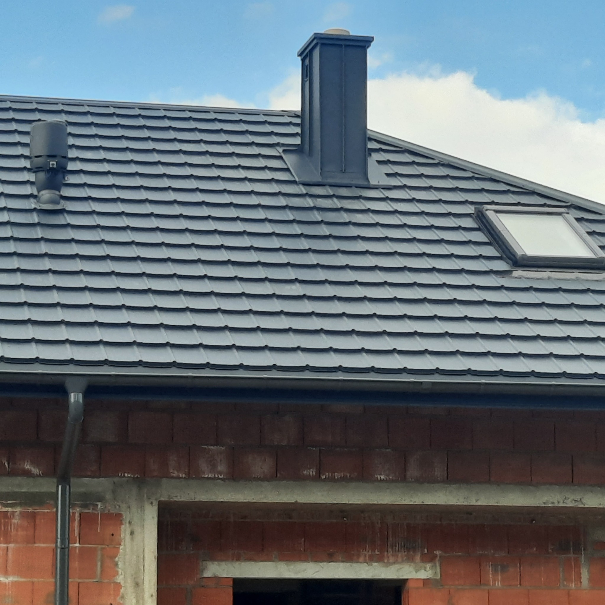 Szary dach z dachówki ceramicznej z kominem, wywietrznikiem i oknem dachowym; widoczna rynna i fragment ściany budynku z czerwonej cegły w stanie surowym.