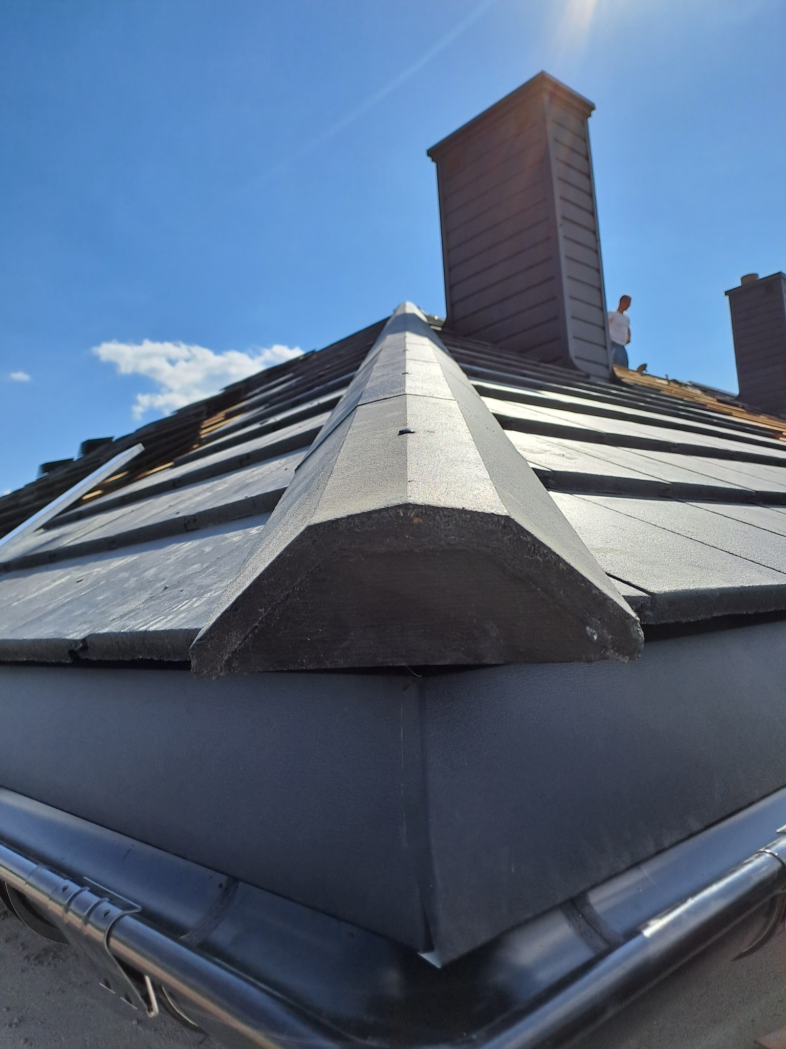 Nowe pokrycie dachu z blachy na rąbek stojący, widok z dołu na krawędź dachu, rynny i komin na tle błękitnego nieba.