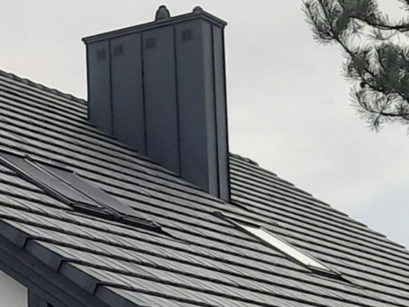 Ciemnoszary dach z dachówki ceramicznej z oknami dachowymi i kominem obłożonym blachą, widok z dołu pod kątem.