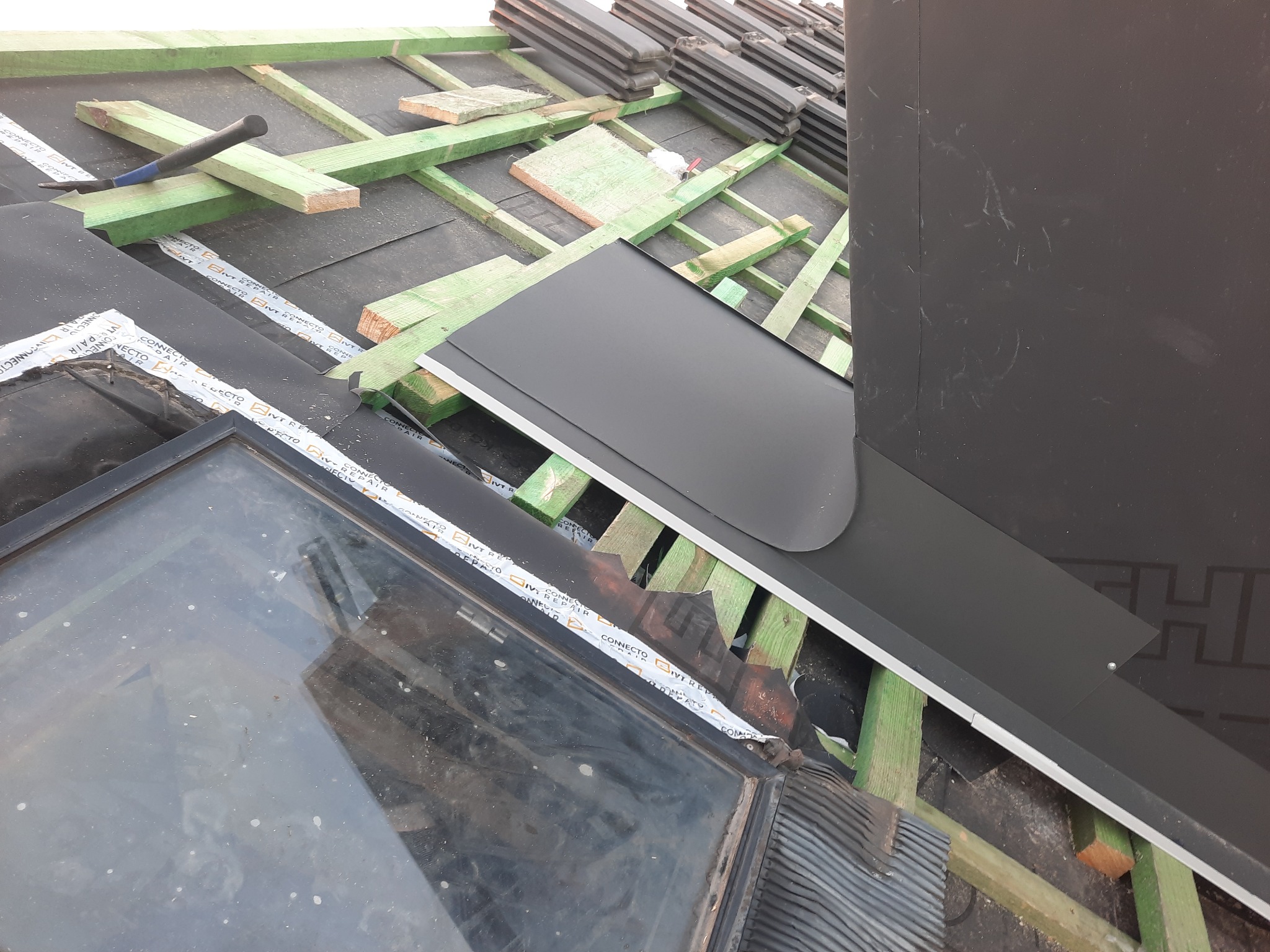 Montaż okna dachowego z czarną obróbką blacharską, widoczne drewniane łaty, fragment dachu pokrytego papą i czekające na ułożenie dachówki ceramiczne, oraz narzędzie dekarskie.