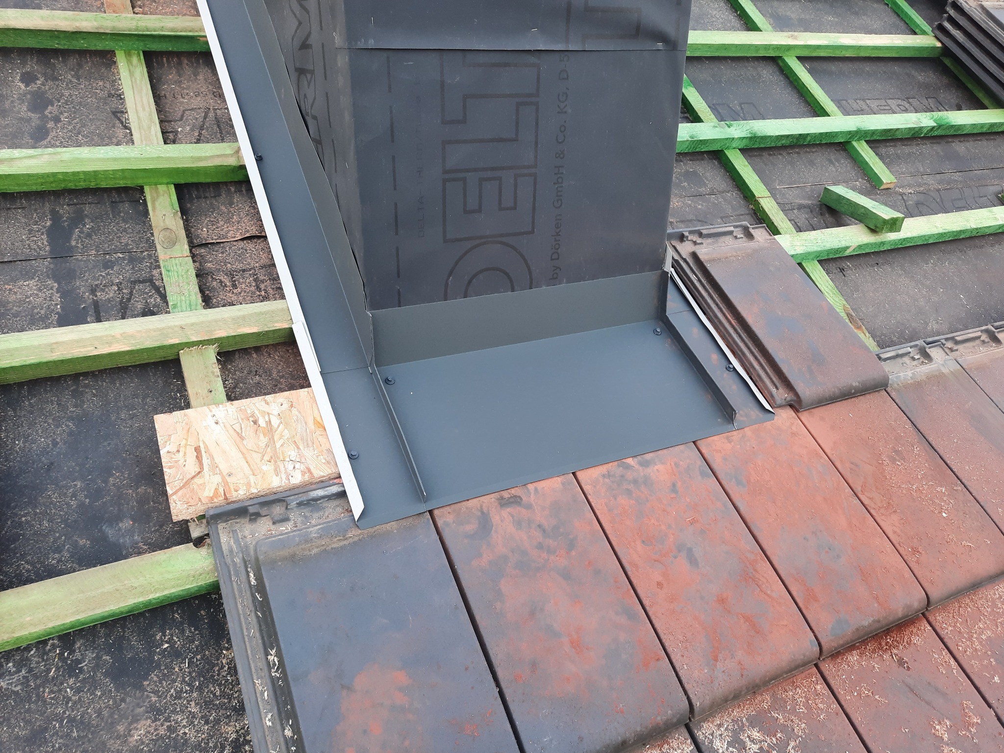 Montaż obróbki blacharskiej komina na dachu pokrytym czerwoną dachówką w Andrespolu, widoczne łaty i kontrłaty oraz membrana dachowa.