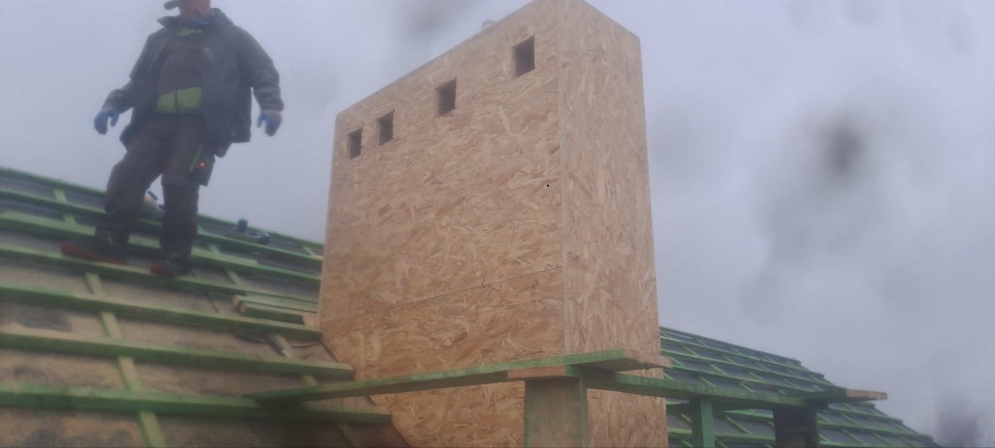 Konstrukcja komina z płyt OSB z czterema otworami wentylacyjnymi w trakcie budowy, widoczny dekarz na dachu z zielonymi łatami w pochmurny dzień.