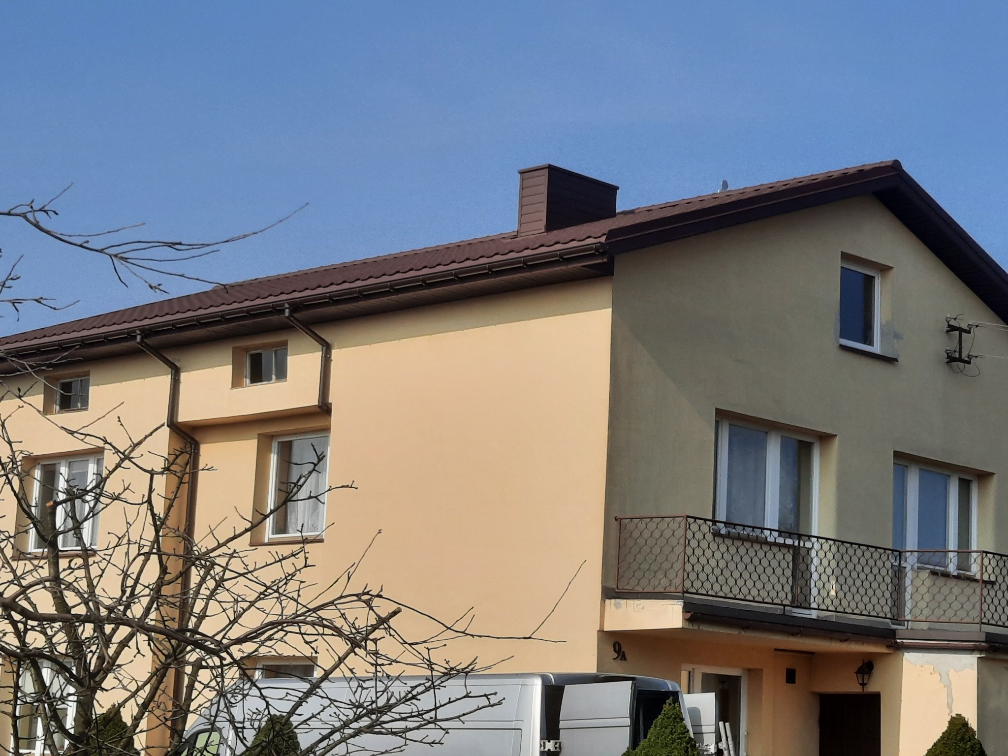 Dwupiętrowy dom z brązowym dachem z blachy trapezowej i brązowym kominem, widoczny z poziomu ulicy z zaparkowanym samochodem dostawczym.