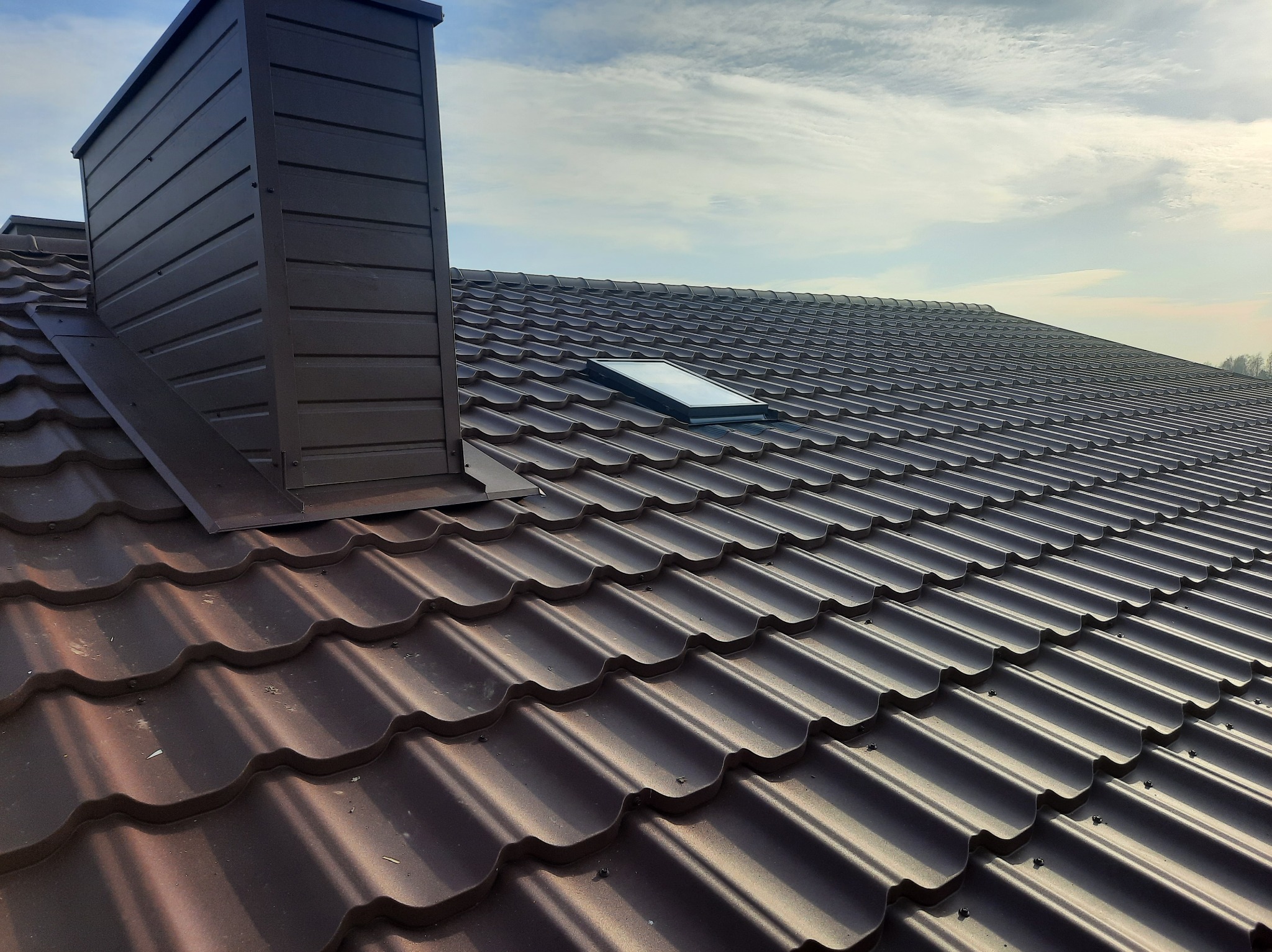 Nowy, brązowy dach z blachodachówki z zamontowanym oknem dachowym i kominem wentylacyjnym, widok z góry na tle błękitnego nieba.