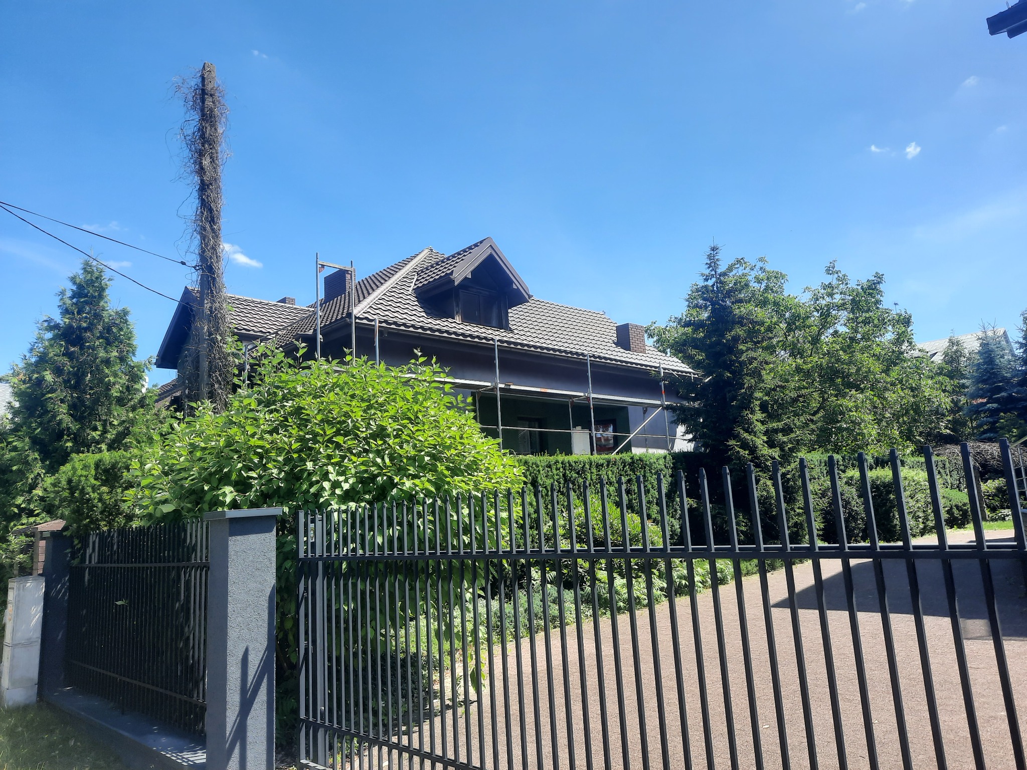 Dom jednorodzinny z ciemną dachówką, częściowo zasłonięty rusztowaniem. Widok zza czarnego, metalowego ogrodzenia w słoneczny dzień.
