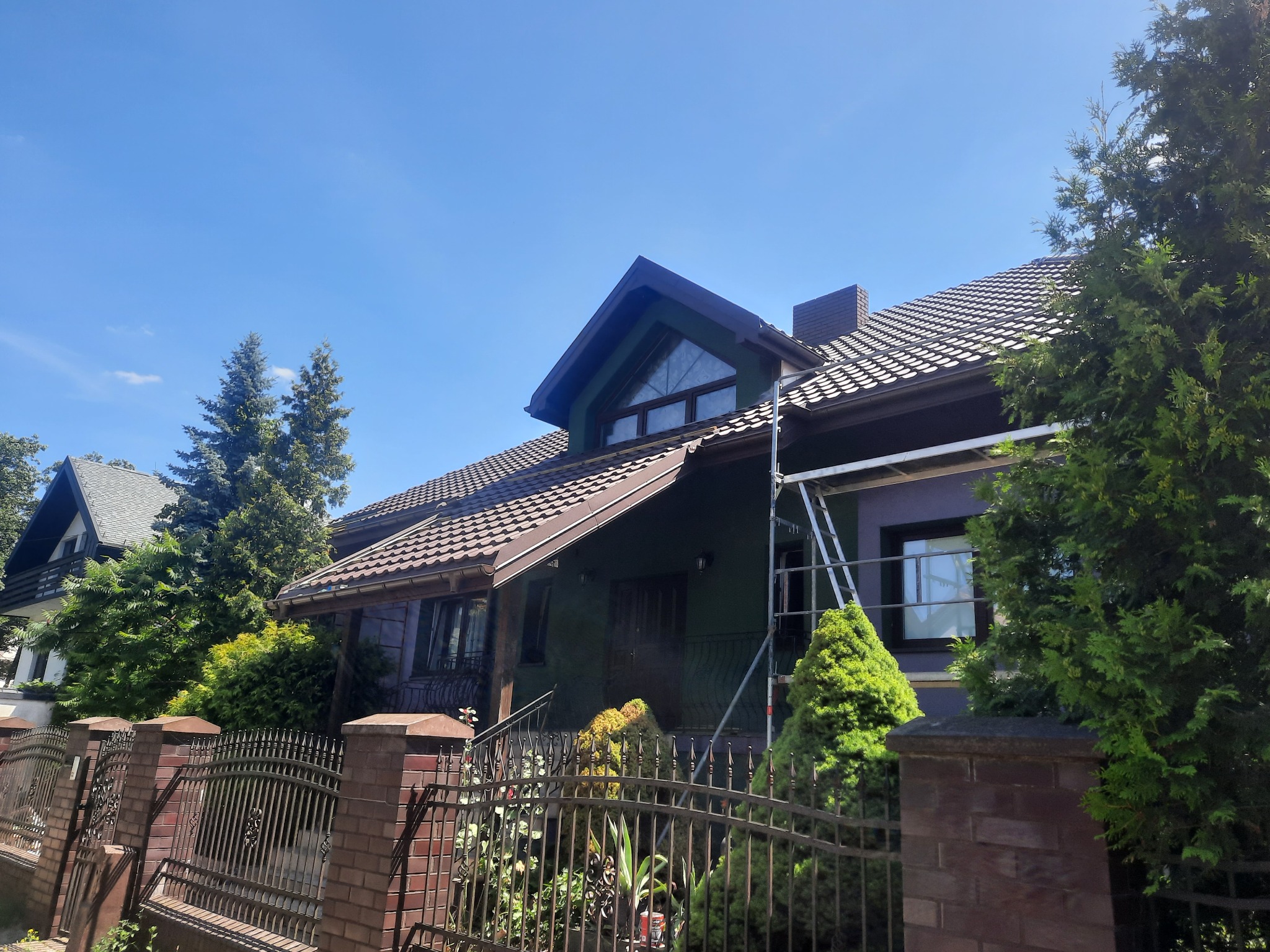 Dom z ciemną dachówką widziany zza ozdobnego ogrodzenia. Na elewacji domu widoczne rusztowanie. Ujęcie w słoneczny dzień.