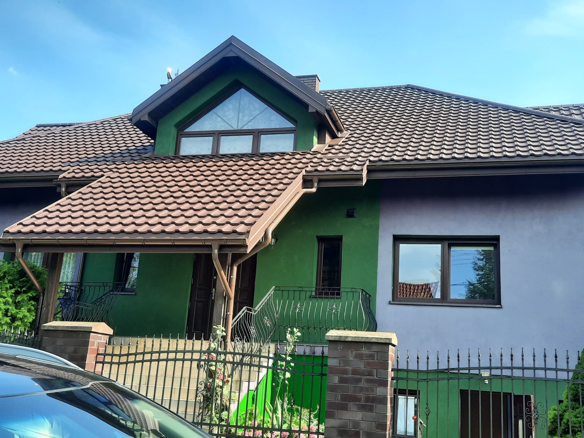 Dom z brązowym dachem z dachówki, zieloną i fioletową elewacją. Widoczne rynny, okna z refleksami i fragment samochodu. Ujęcie z dołu, w słoneczny dzień.