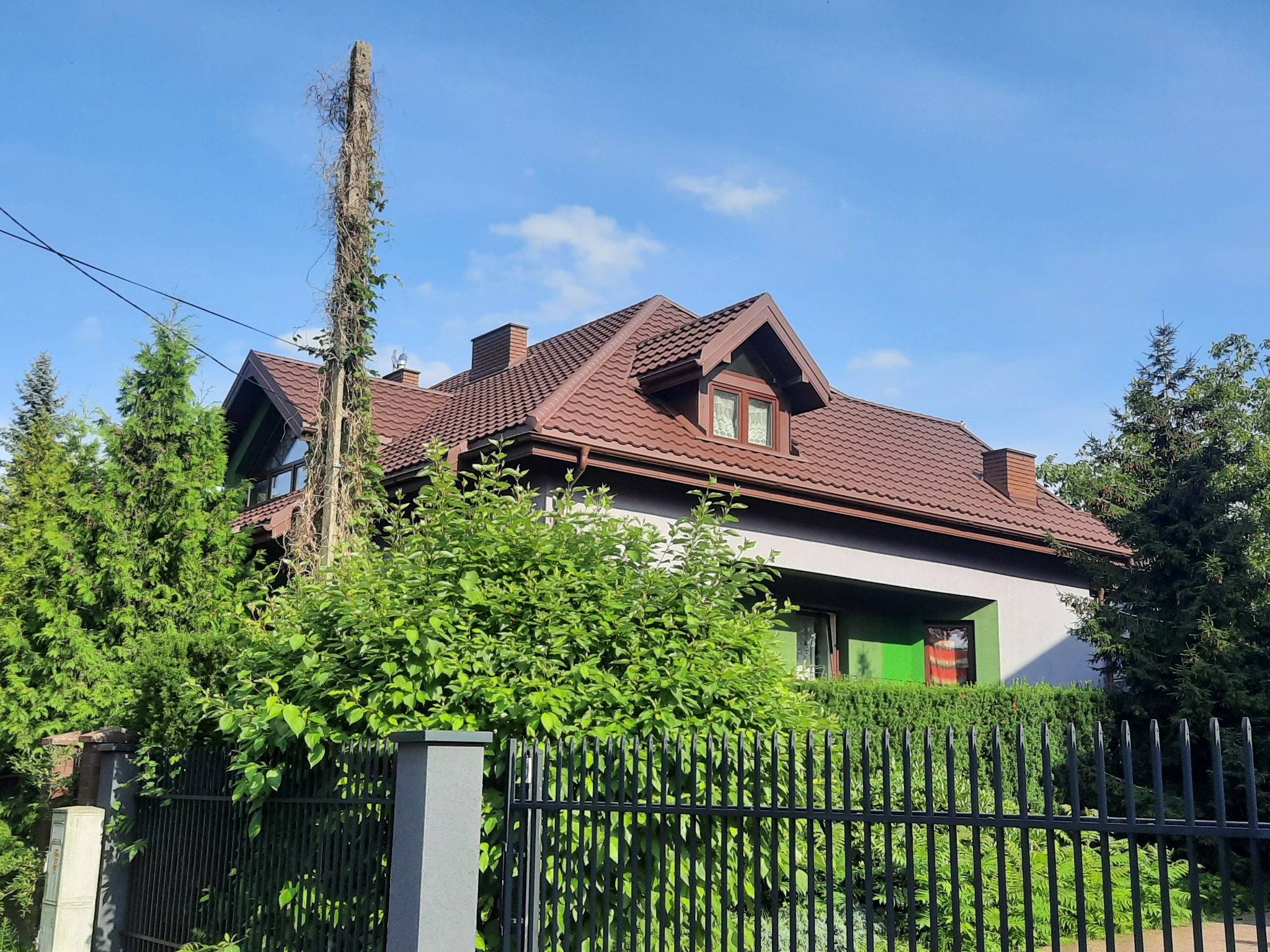 Dom jednorodzinny z brązowym dachem z blachodachówki, widoczny zza czarnego ogrodzenia i zieleni. Na dachu kominy i okno dachowe.
