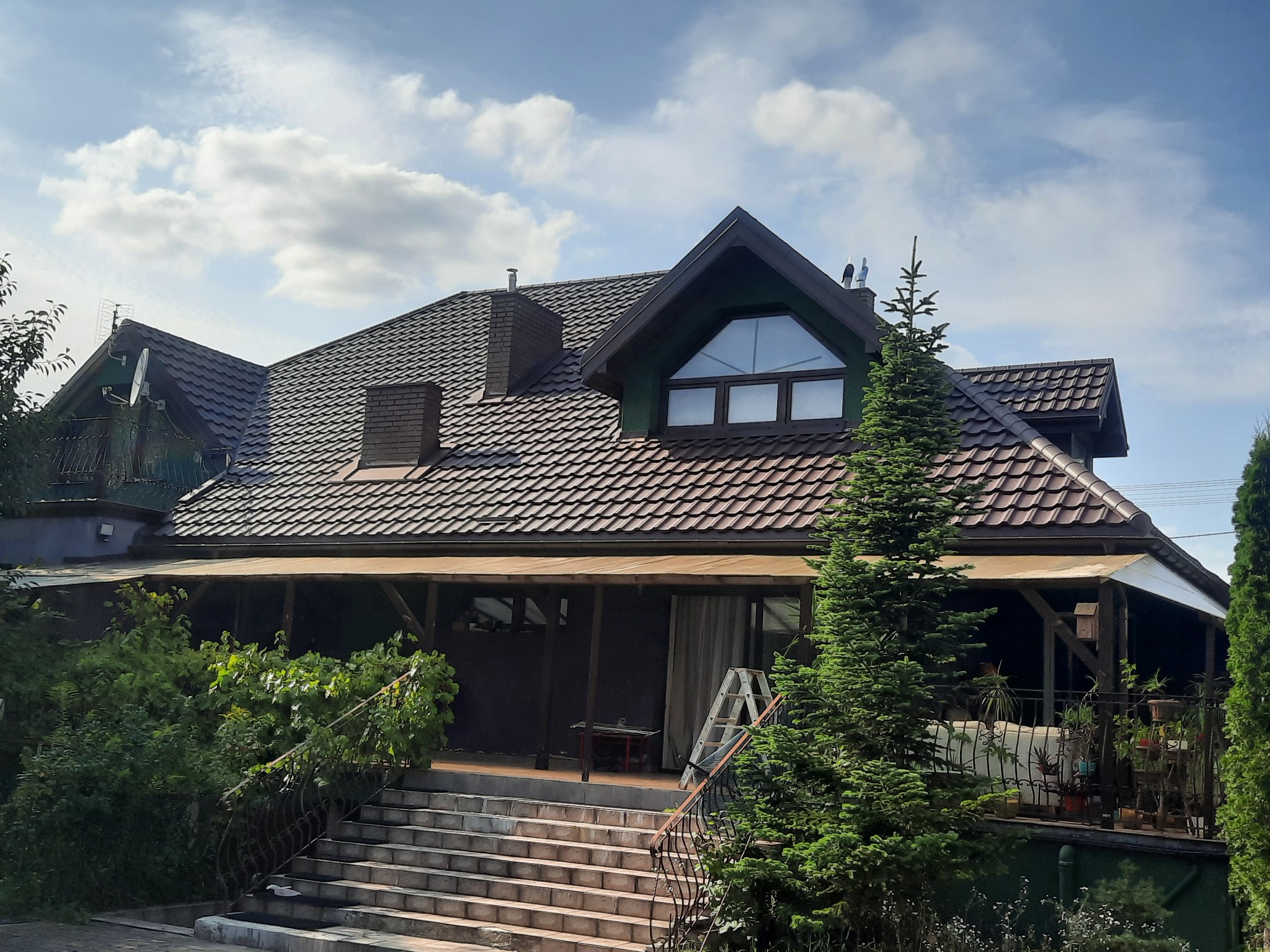 Dom jednorodzinny z brązowym dachem z dachówki, widoczne schody wejściowe, fragment ogrodu i drabina oparta o ścianę. Ujęcie w słoneczny dzień.