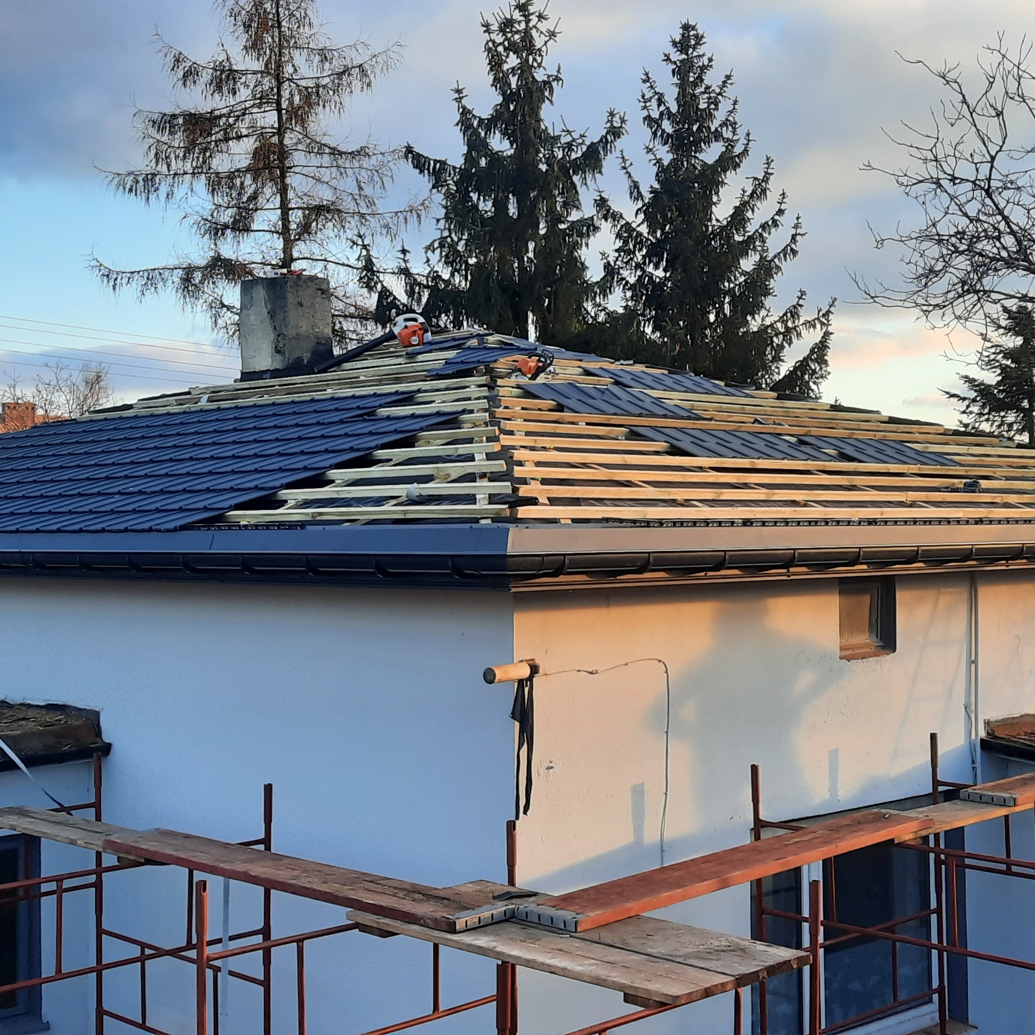 Dom w trakcie krycia dachu blachodachówką. Widoczne rusztowania, drewniana konstrukcja dachu i częściowo ułożone panele. Na dachu leży piła i kask.