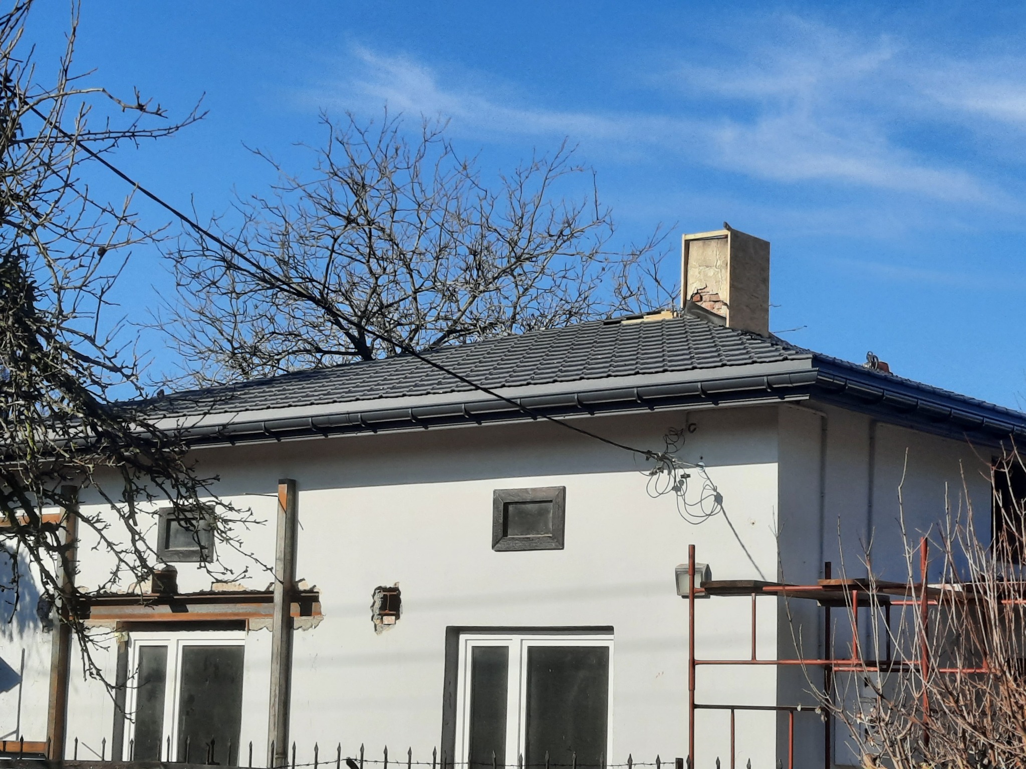 Szary dach z dachówki ceramicznej na białym domu jednorodzinnym, widok z dołu na tle błękitnego nieba. Widoczne rusztowania i elementy przygotowania do prac remontowych.