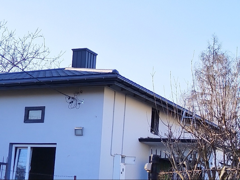 Dom jednorodzinny z ciemnym dachem i kominem w słoneczny dzień. Widoczne rynny i obróbki blacharskie. Na pierwszym planie gałęzie drzew.