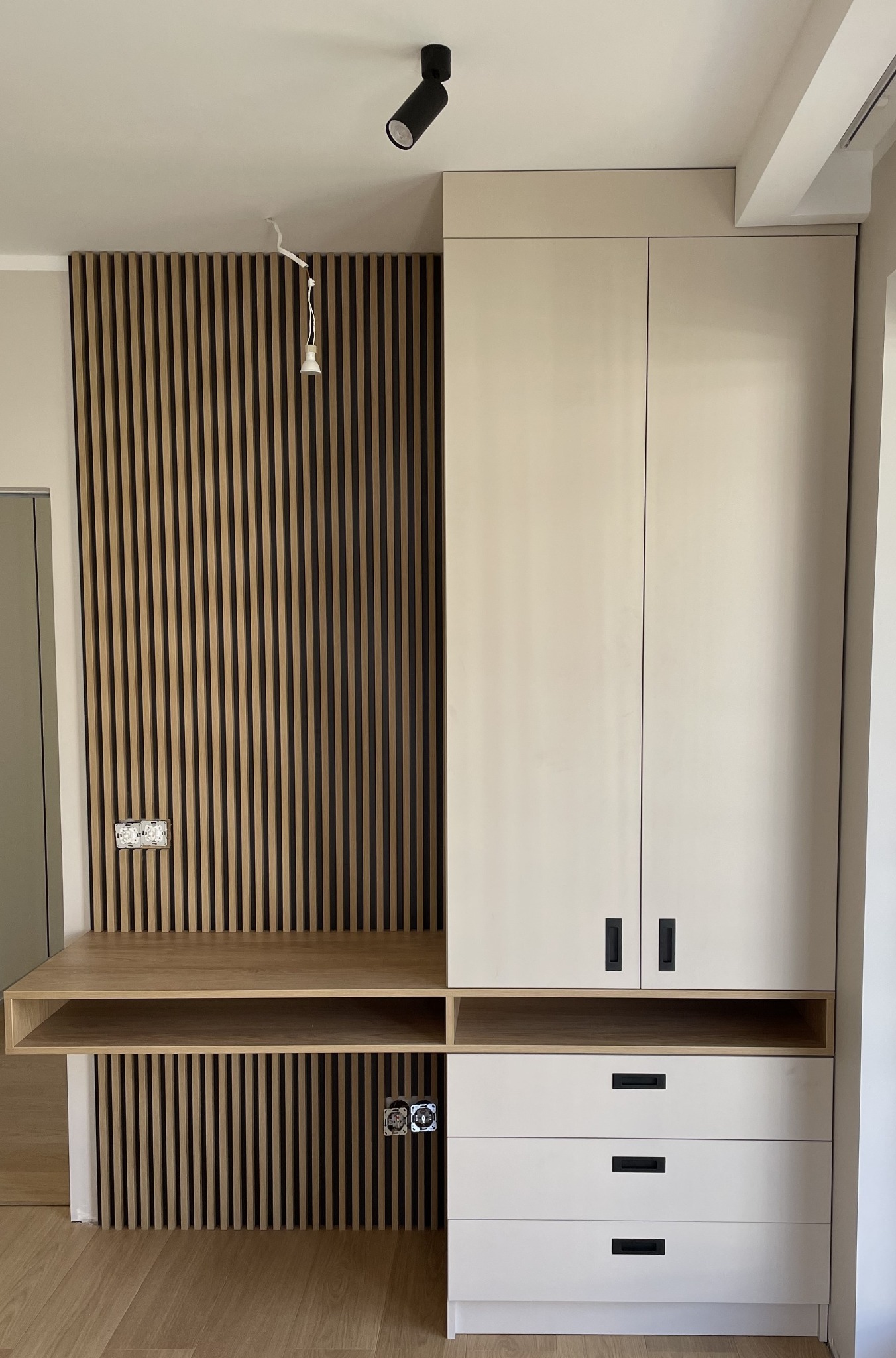 Nowoczesna szafa na wymiar z drewnianymi lamelami na ścianie, jasne fronty, czarne uchwyty, biurko wkomponowane w szafę, widoczne gniazdka elektryczne na ścianie z lamelami.