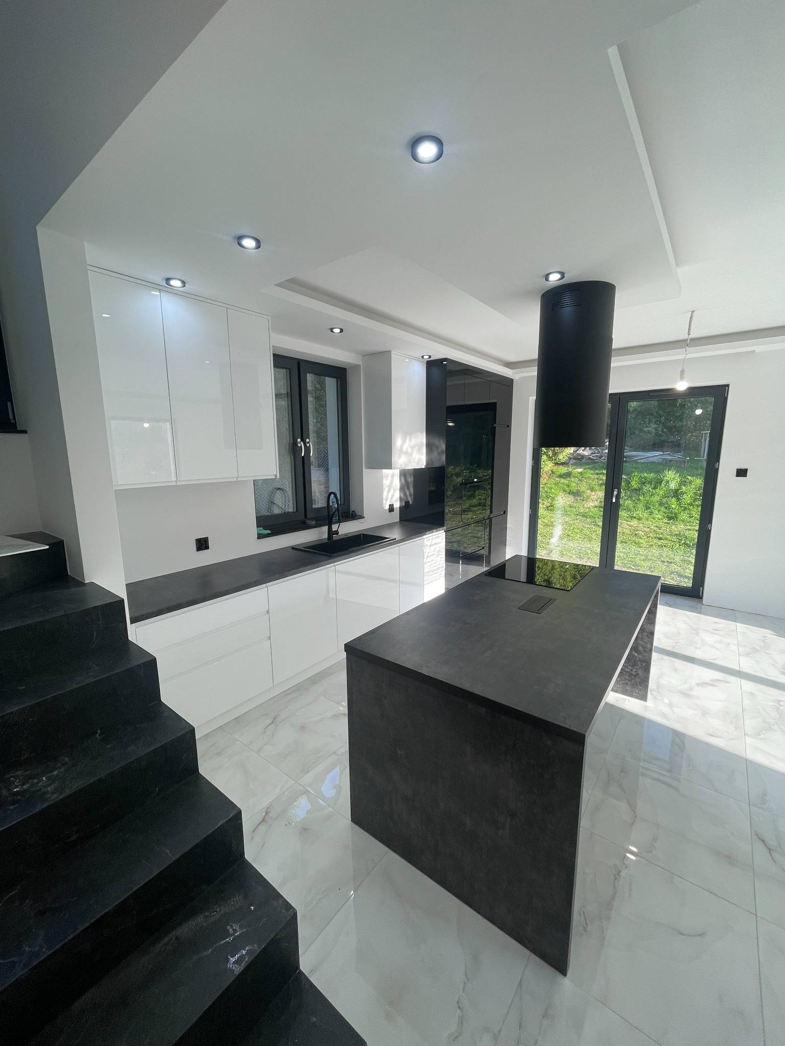 Nowoczesna, jasna kuchnia z białymi szafkami, czarnym blatem i wyspą kuchenną z czarnym okapem, widok z dołu schodów.
