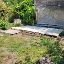 Brukisiema Mateusz Szczepaniak - Przygotowany teren pod wylewkę, widoczne resztki starej konstrukcji, zarośnięty trawnik i fragment ściany budynku w tle. Prace ziemne przed wylaniem fundamentów.