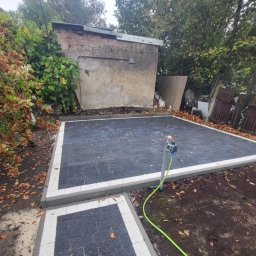 Brukisiema Mateusz Szczepaniak - Ułożona, ciemnoszara kostka brukowa z białym obramowaniem, tworząca plac. W tle budynek gospodarczy i zieleń. Widoczny kran z podłączonym wężem ogrodowym.